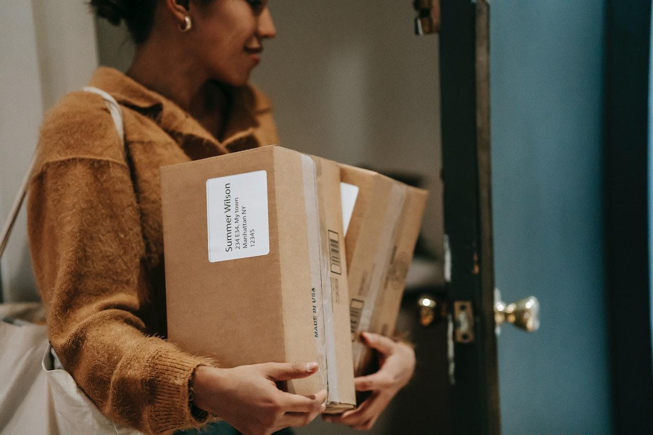 A young woman wearing a yellow jumper carries two rectangular boxes through a blue door